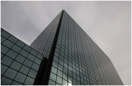 A view of a glass building’s facade from the angle of a pedestrian on the street. The building’s facade is a grid of glass and otherwise without defining features.