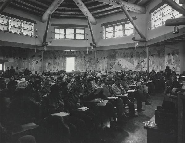 A large crowd, men and women, sit in a circular room decorated with murals and a clerestory, styled as a hogan.