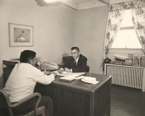 Two men sit across an office desk from one another; one, smiling, takes notes. A painting of a Navajo man hangs on the wall.