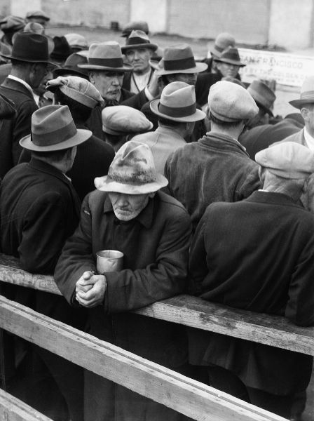 A crowd of men, most with their backs to the camera, but one, facing, leans on a fence rail, cradling a metal cup with his forearms.