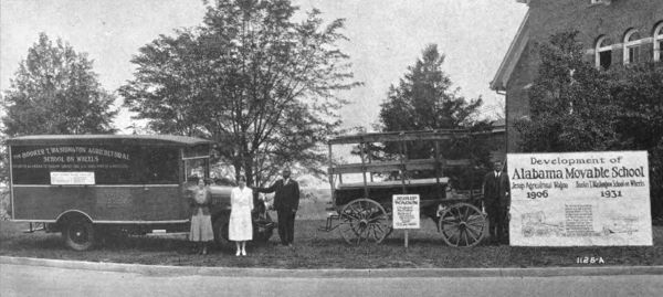 Grainy photograph showing three people standing beside an early truck with a large covered flatbed. Next to it a wagon with wooden wheels that could be pulled by a team of horses.