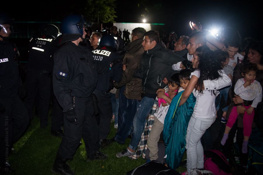 Roma families are attacked in the night by police. In the middle of the photo, a family is holding two small children close. A teenager is standing in front of them blocking a police officer.