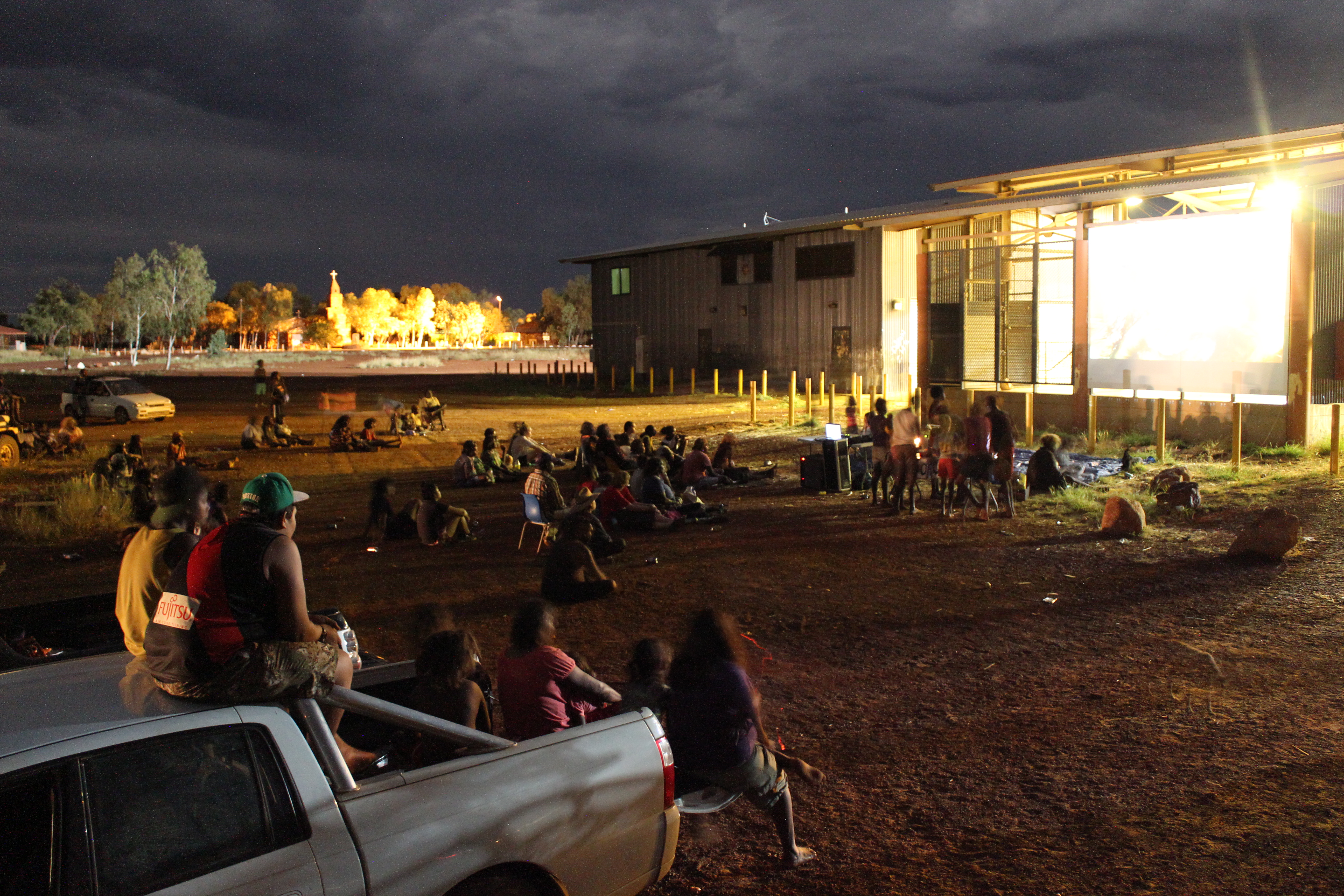 In this long-exposure night picture, dozens of audience members sit on the ground or on trucks in a flat pebbled area, facing a bright cinema screen.
