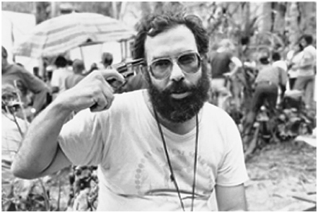 Francis Ford Coppola poses for a picture holding a revolver to his temple. He’s wearing a t-shirt, aviators sunglasses, and has a full beard. Film crew are visible in the background.