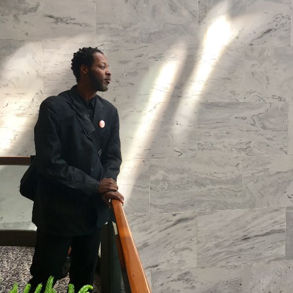 Indoor shot of a man in black with a slight smile looking out over a wooden railing. Behind him, two sunbeams shine on a marble wall.