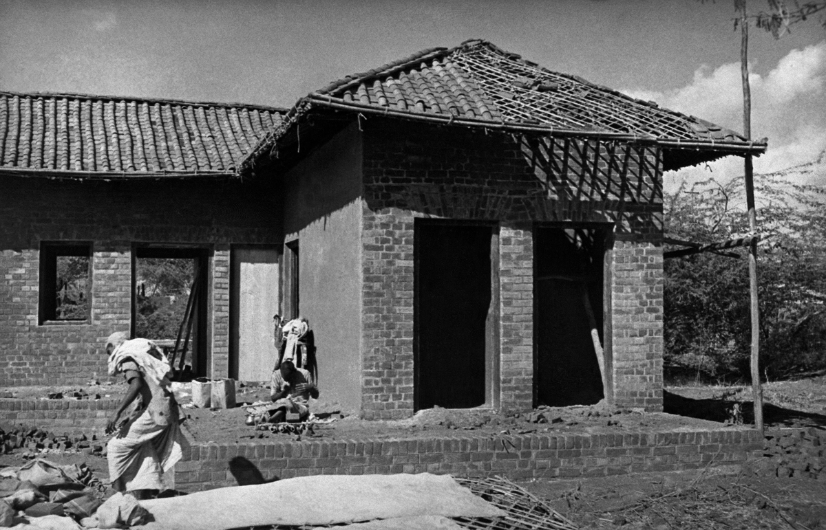 Photograph showing brick building under construction with unfinished roof of ceramic tiles and two workers.