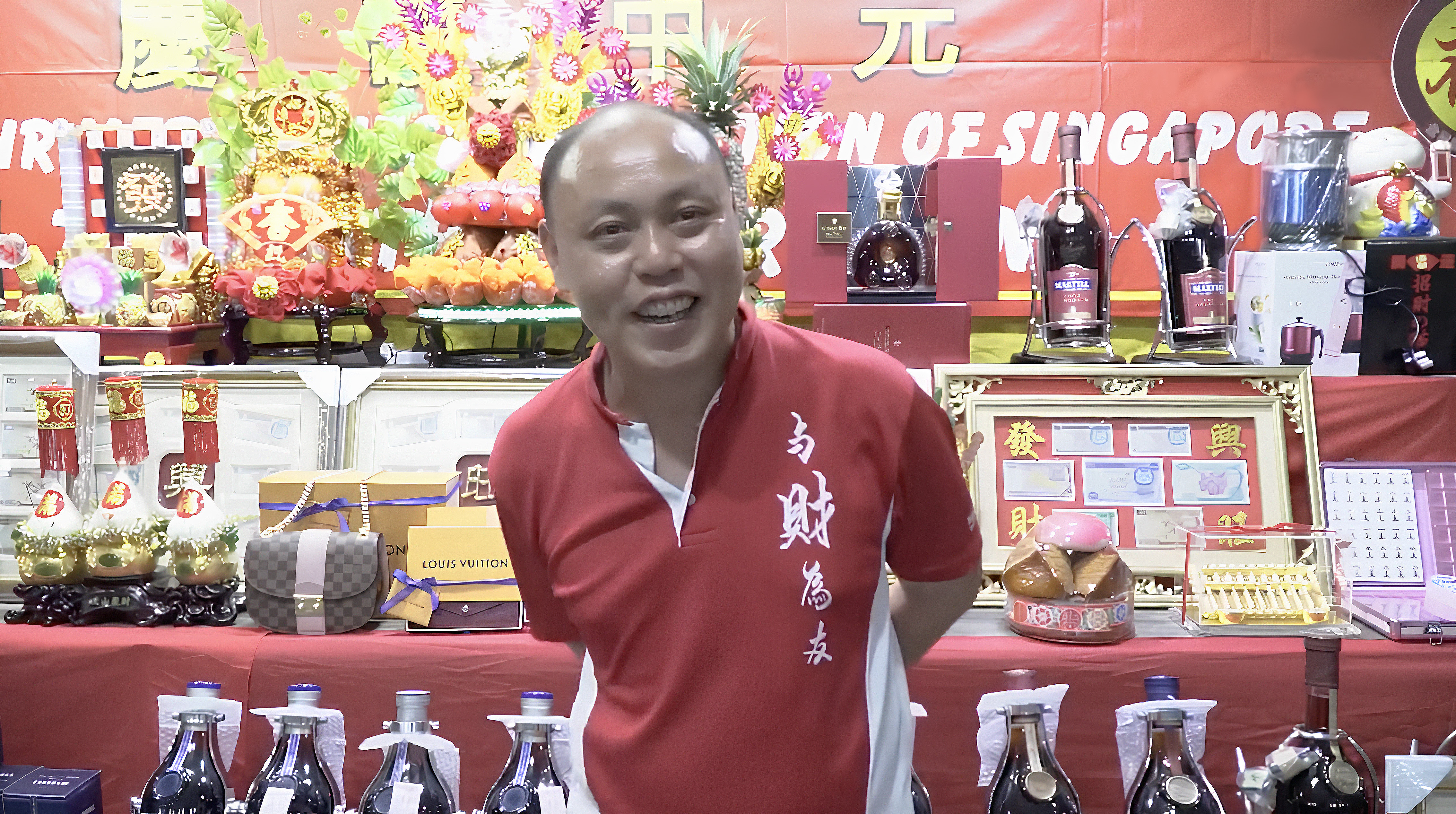 A smiling man in a red shirt stands in front of tiers of goods on a dais. These include liquor bottles, religious icons, and luxury items.