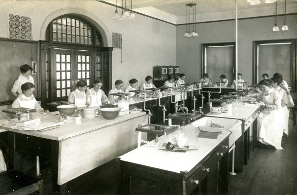 A group of women, each wearing a white apron, seated next to one another around a large U-shaped table equipped with plumbing.