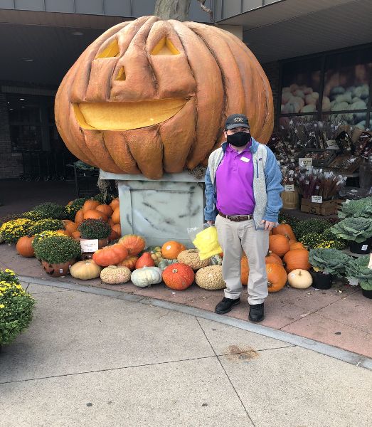 Jake beside a Halloween pumpkin display at his job. He wears tan pants, a Wegmans shirt, hat, and name tag, a gray and blue jacket, and a black mask. 