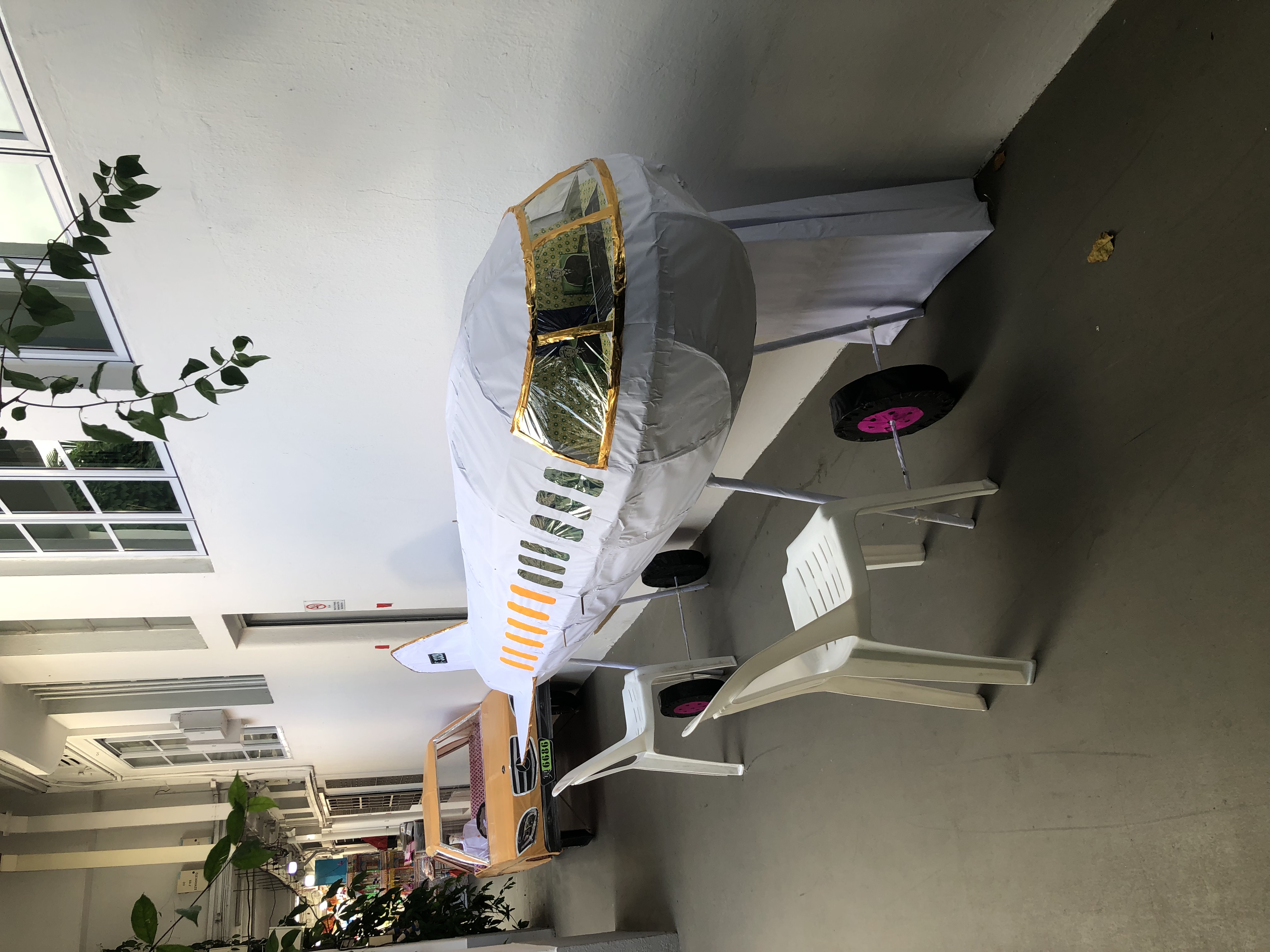A paper car and paper passenger plane stand at the wall of a building. These stand several feet tall, and include drivers, pilots, and other detail.