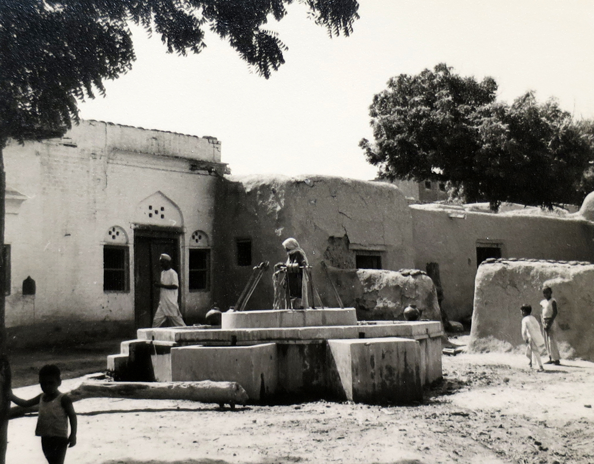 Photograph of village well with several children and adults, one standing on the well and the others walking by.