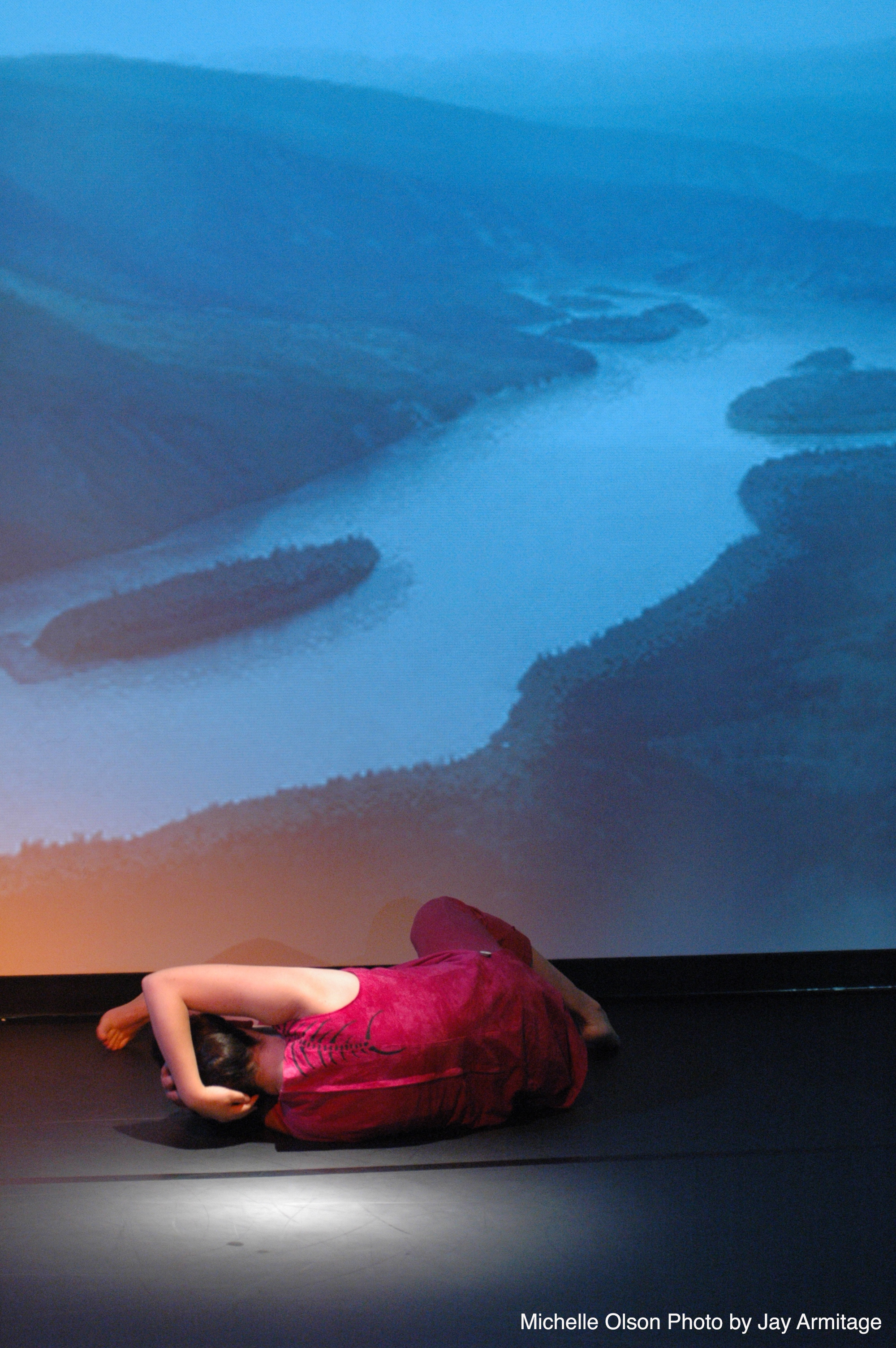 Performer on stage in fetal position, facing toward a backdrop onto which an image of a valley and river are projected.