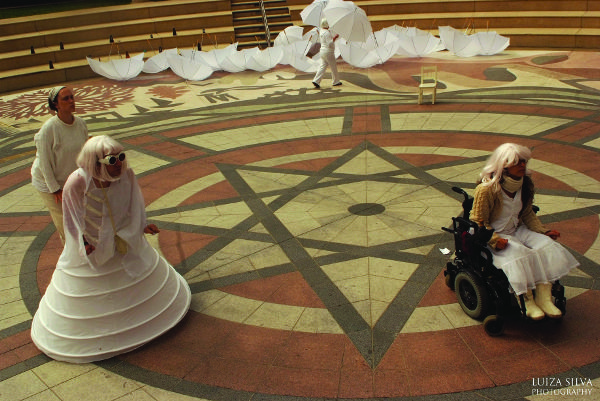 Four figures dressed in white in a plaza; the one in the background holds a white umbrella; one figure in the foreground is in a wheelchair.