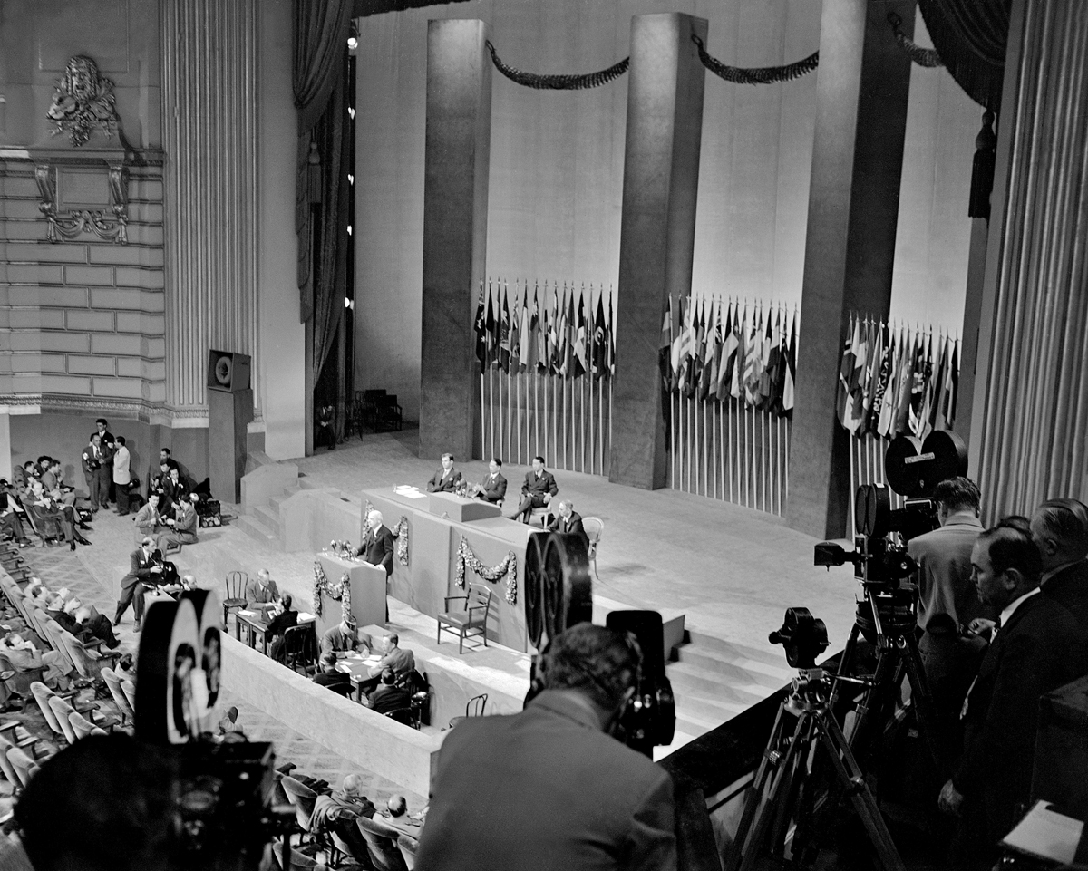 Photograph of men with cameras on a gallery filming a speaker behind the podium on the stage, with pylons and flags forming the background.