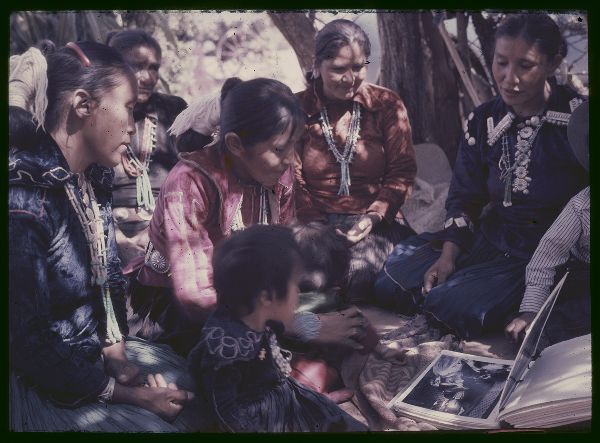 Six smiling women and children seated in a traditional summer shelter look at a book of photographs on the floor between them.