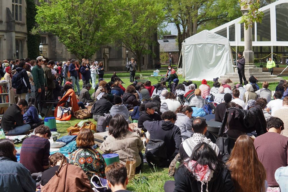 Students, faculty, and community members at Princeton University sit on the grass listening to a speaker. Security personnel are seen in the distance.