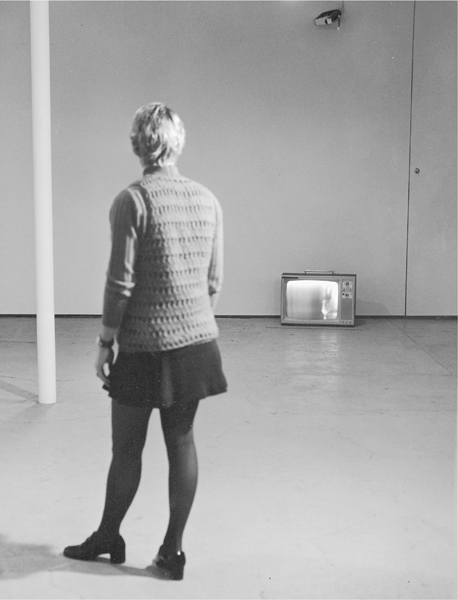 A photograph of a woman standing in a room looking at a television placed on the floor at the far end.