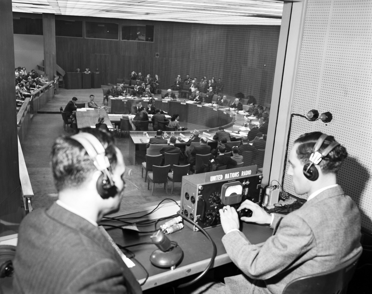 Photograph of broadcasting booth with two men in headphones looking at delegates convening around a semicircular table.