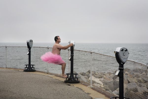 A man, dressed only in a pink tutu, standing on the foot of a coin-operated bincoulars looking out over a large body of water.