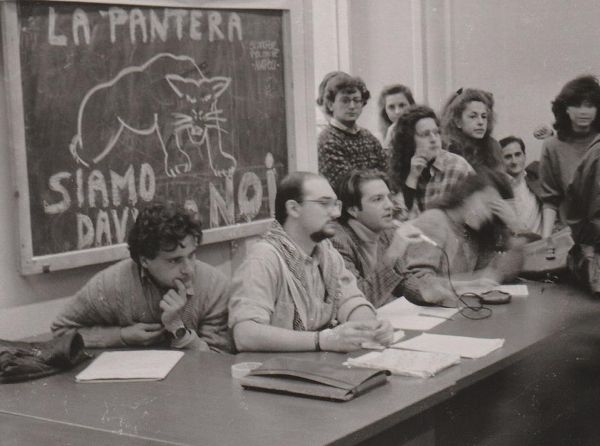 Black-and-white photo of four students sitting behind a desk, speaking with a microphone to an assembled group not pictured; in the background a chalkboard with a drawing of a panther beneath the heading “La Pantera.”