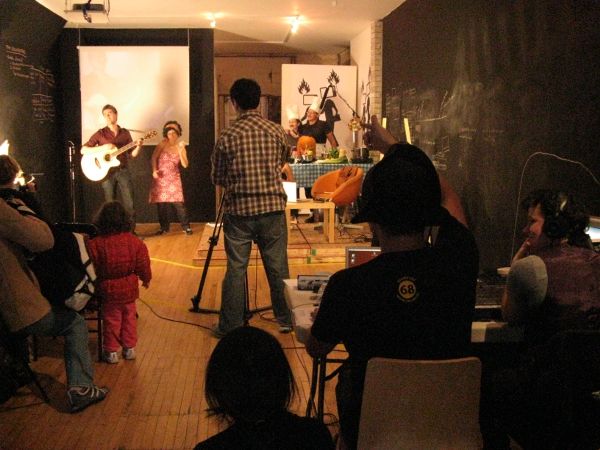 A behind-the-scenes shot depicting a person working a camera, filming a live-music segment; off to the side are participants for a home-cooking segment. In the foreground, people watch on from plastic chairs.