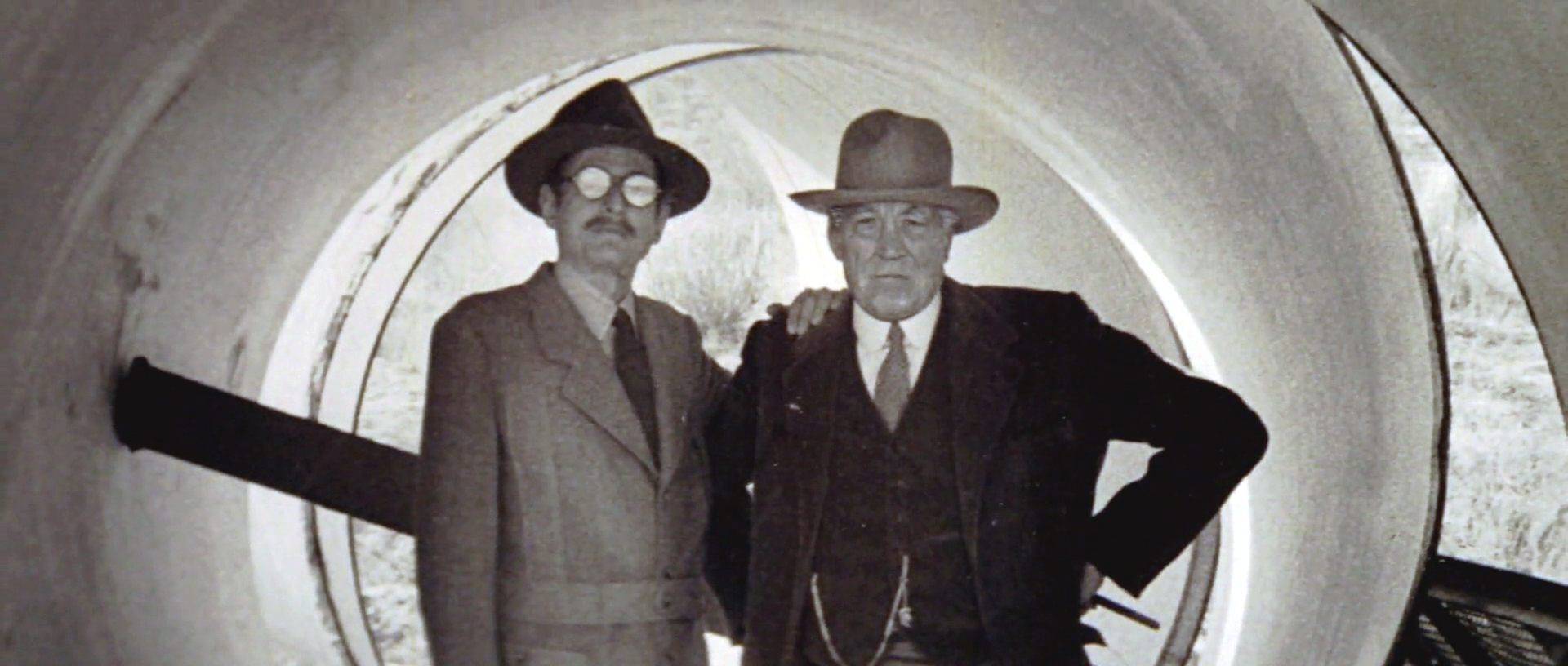 Two well-dressed men stand together inside of an 8-foot diameter pipeline.