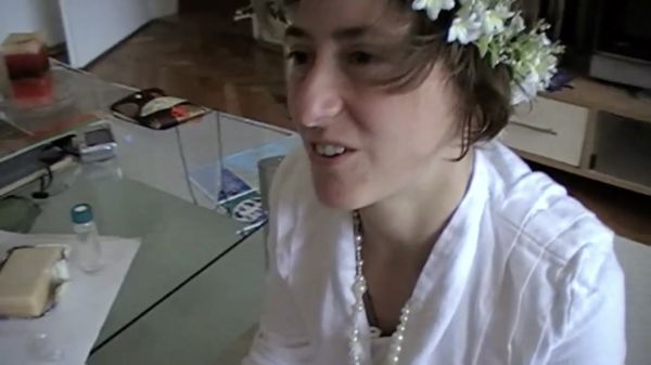 A woman in white with a flower tiara kneeling on the ground with a smile on her face.