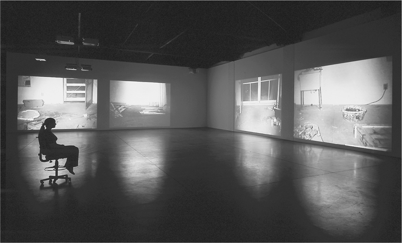 A photograph of a big room with 4 projectors, 2 on each wall. A woman is seated on a chair facing a wall with 2 projectors. All the projectors have pictures on them.
