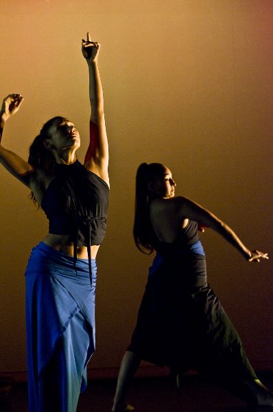 Two women in long skirts and short tops stand. One looks up, slightly arching, arm lifted. The other, in profile, shifts hips to side, arm curving.