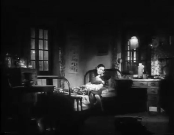 Dark image showing the interior of a bedroom with a figure on the bed clutching something to their chest.