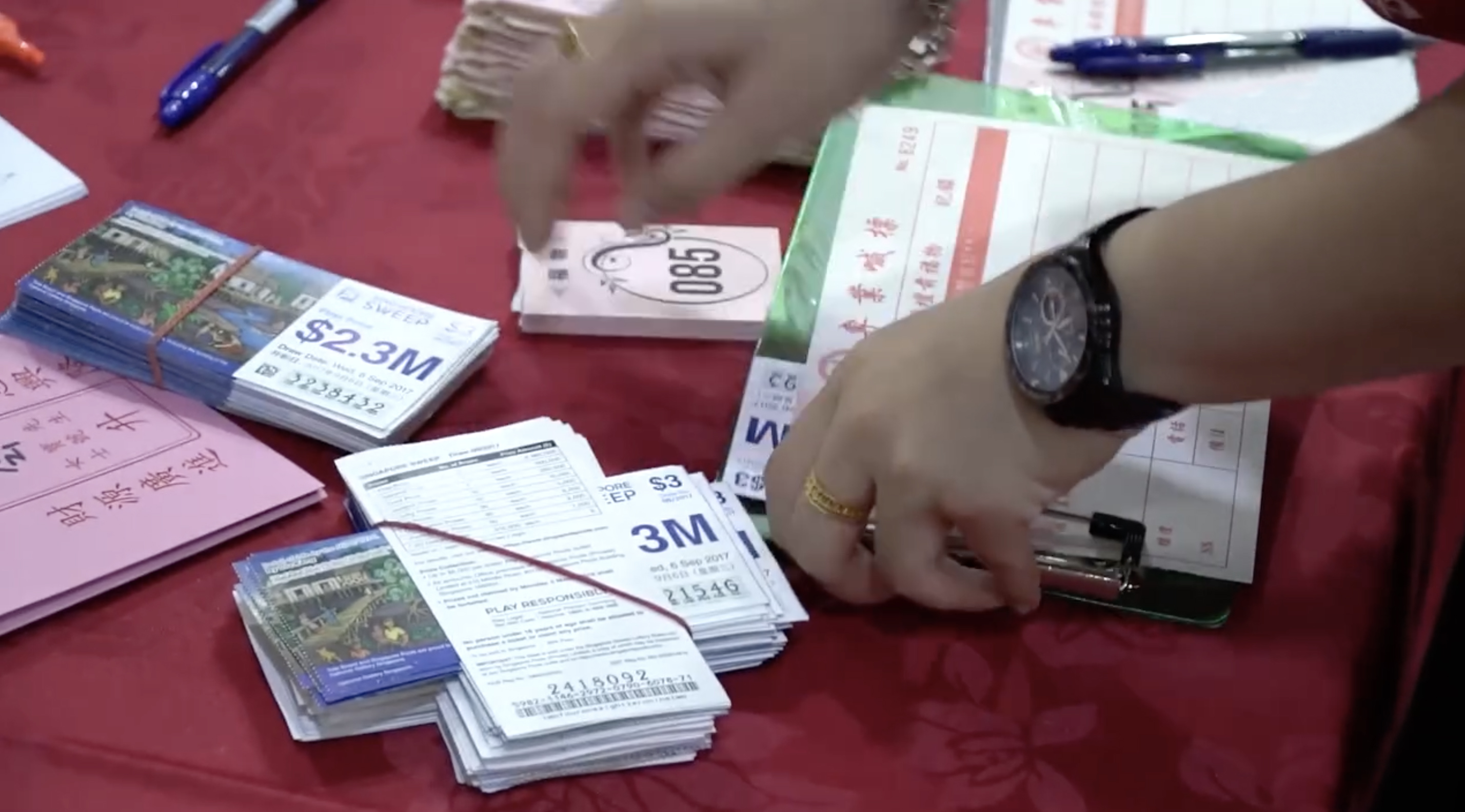 A pair of hands are arranging neat stacks of paper on a red table. These are lottery tickets, with a prize of 2.3 million Singapore dollars.