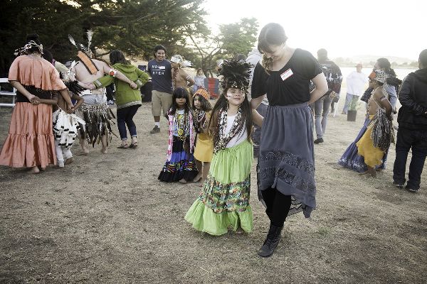 Couples dance with arms crossed behind them. In the front, a girl in a long skirt and feather headdress dances with a smiling woman with a name tag.