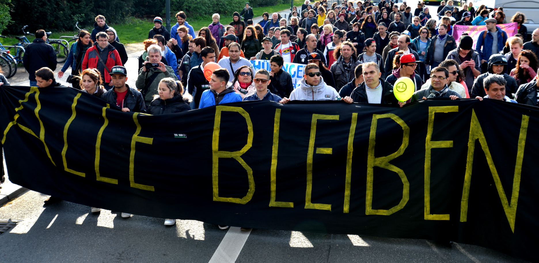 Several people are holding up a black banner with the words “ALLE-BLEIBEN!” (all stay) in yellow. Behind them are dozens of people walking in solidarity against Roma deportations.