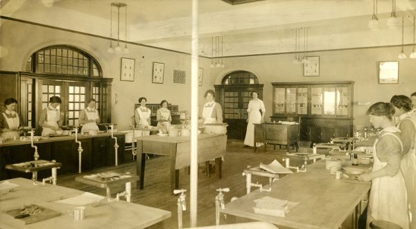 A large room with several work tables equipped with plumbing. Around ten women are standing at various stations working. 
