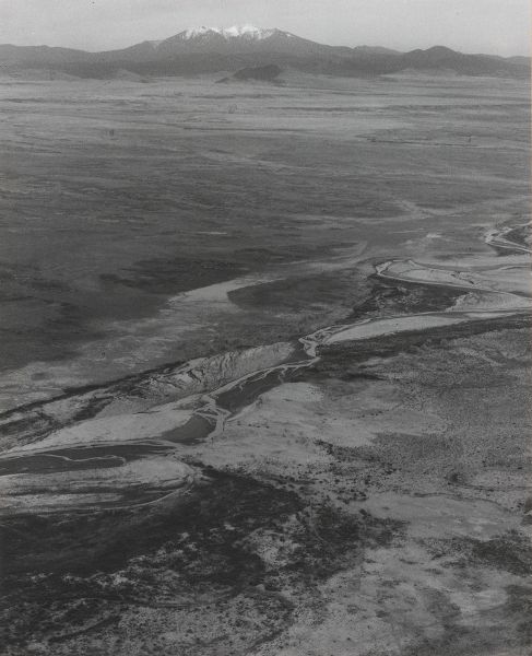 An aerial view of a river in the foreground and a single snow-capped peak among smaller peaks on the horizon.