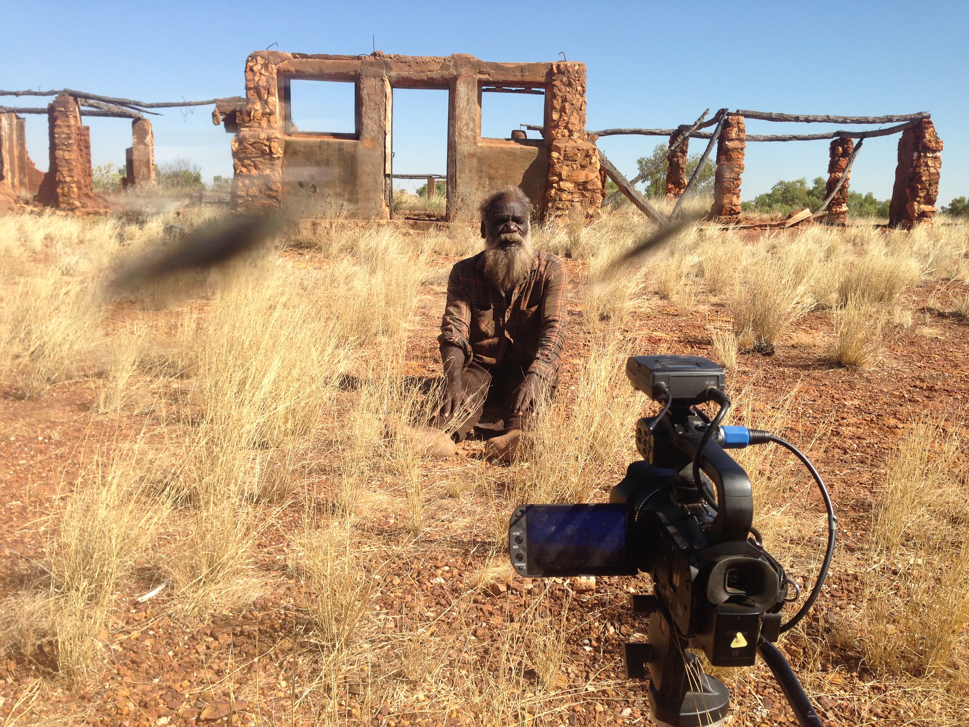 Background shows the brick and stick remains of Old Balgo. Mark sits on the ground amid desert grasses, looking directly into the camera.