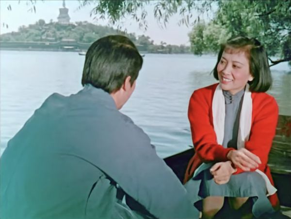 A young man and woman sit facing one another in a small boat; the woman wears a bright orange sweater. A monument appears in the distance.