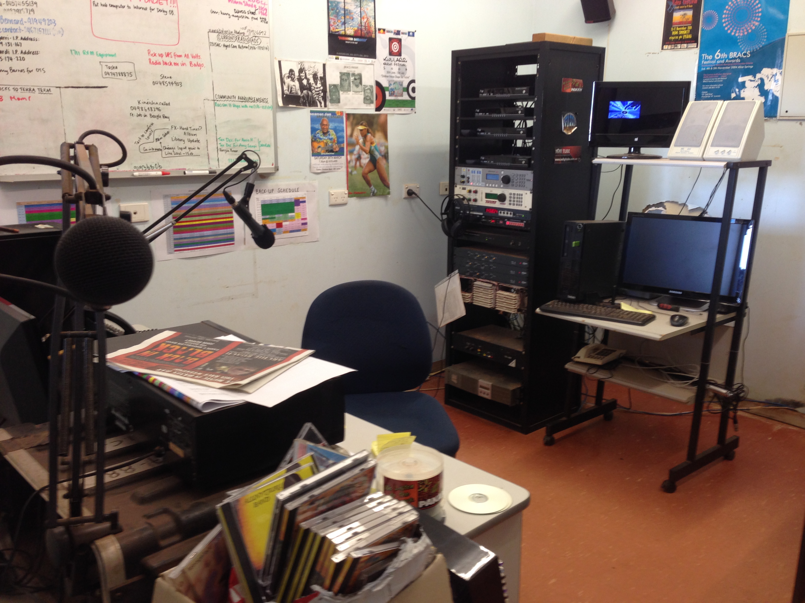A rolling chair in the middle of the room sits in front of a radio desk. Behind it are electronic equipment, another computer, and a whiteboard.