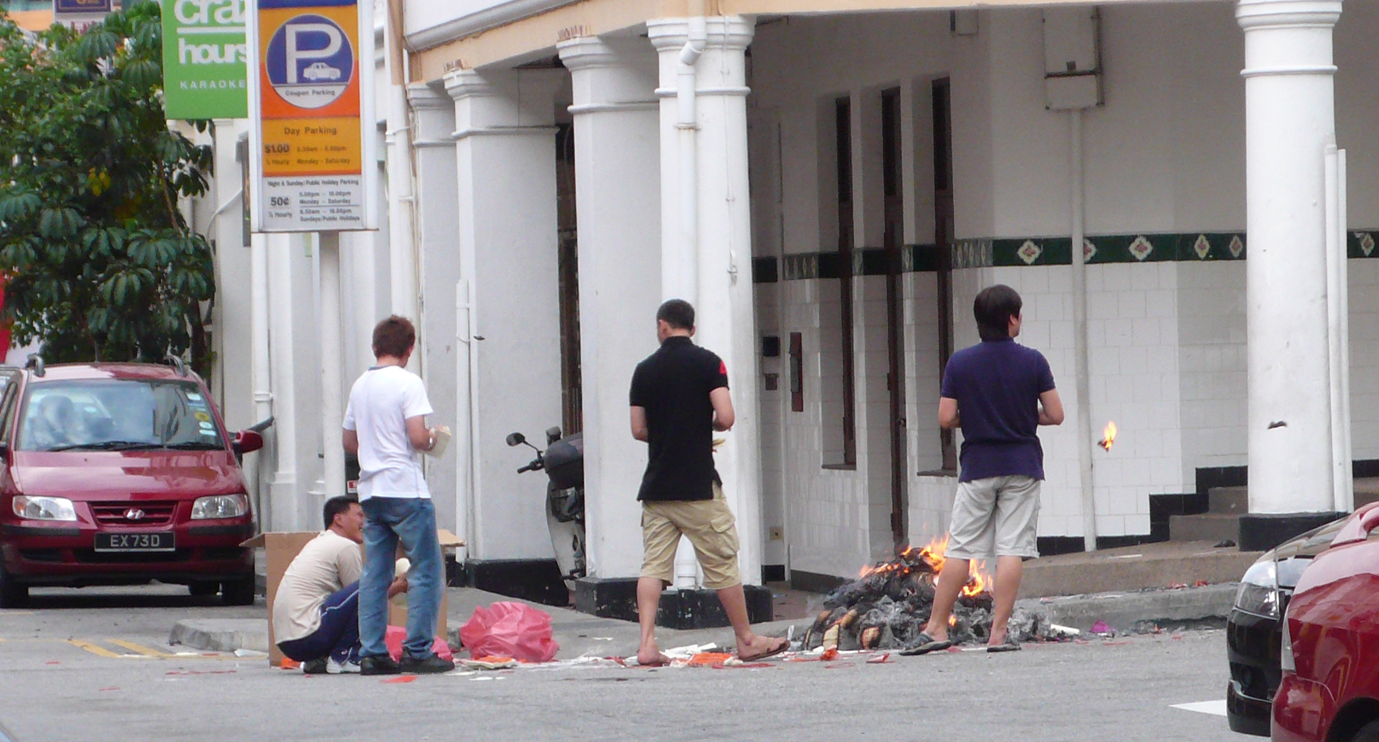 Four men stand around a fire on a streetcorner, surrounded by colonial buildings. Between them is a large pile of smoldering ash and refuse.