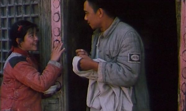 A woman in a red coat and a man in a gray coat stand beside a doorway speaking to one another.