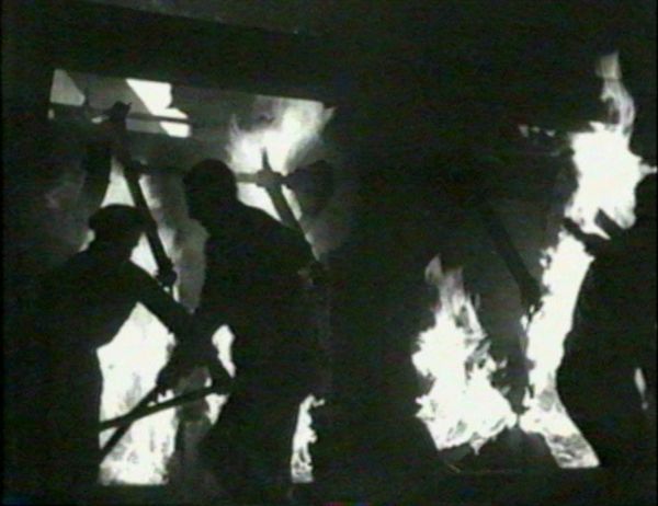 Three men in shadow work with and melt steel as large flames engulf the factory scene behind them.