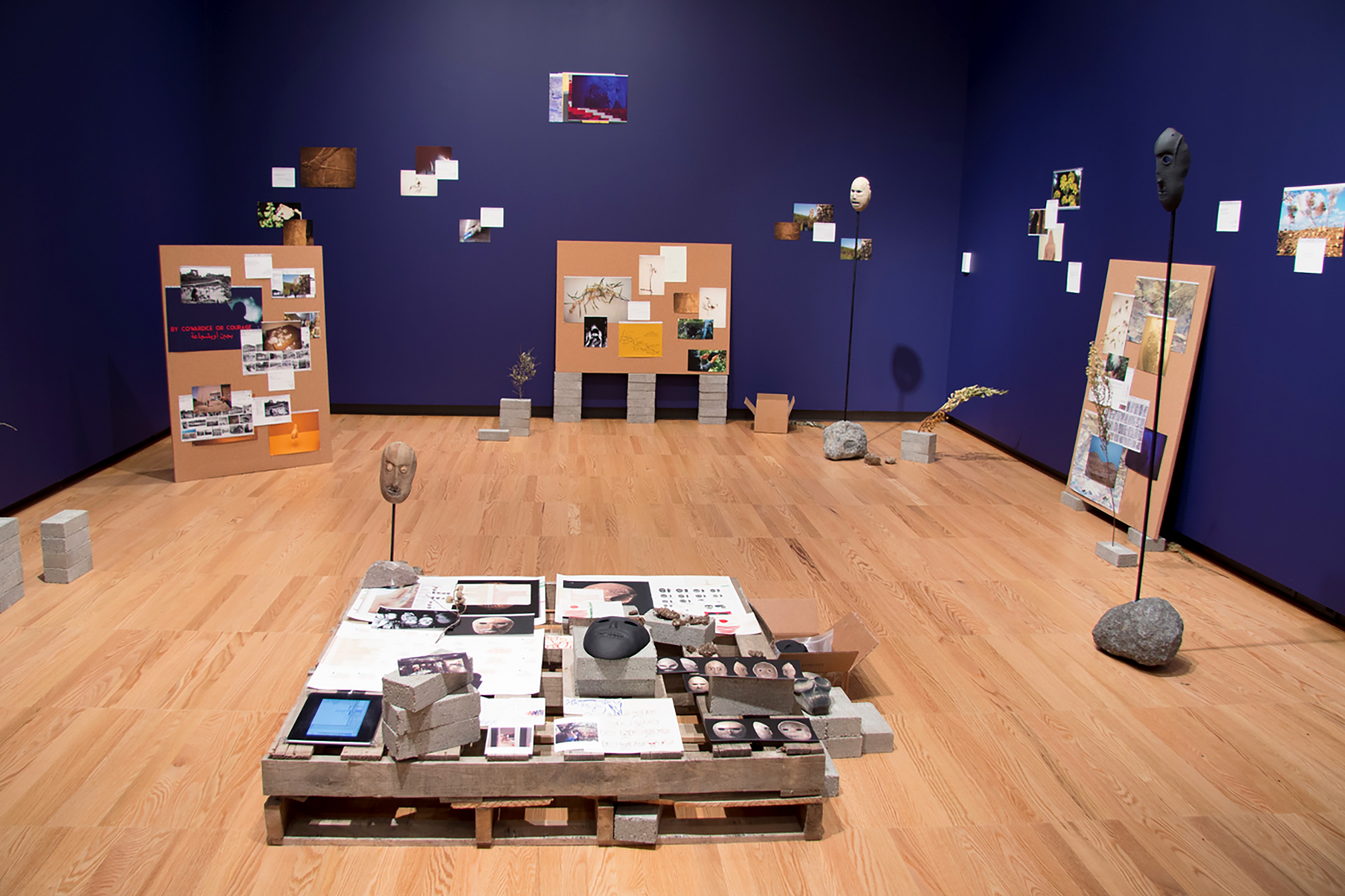 A large room with wood flooring and blue walls populated with various artifacts: makes on piles, drawings and pictures on the walls and boards, a pallet with the same upon it. Stones and gray bricks accent throughout.