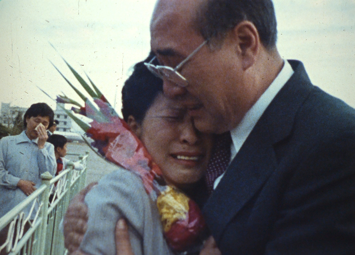 An older, balding man in a suit and tie embraces a woman, crushing a bouquet of flowers between them.