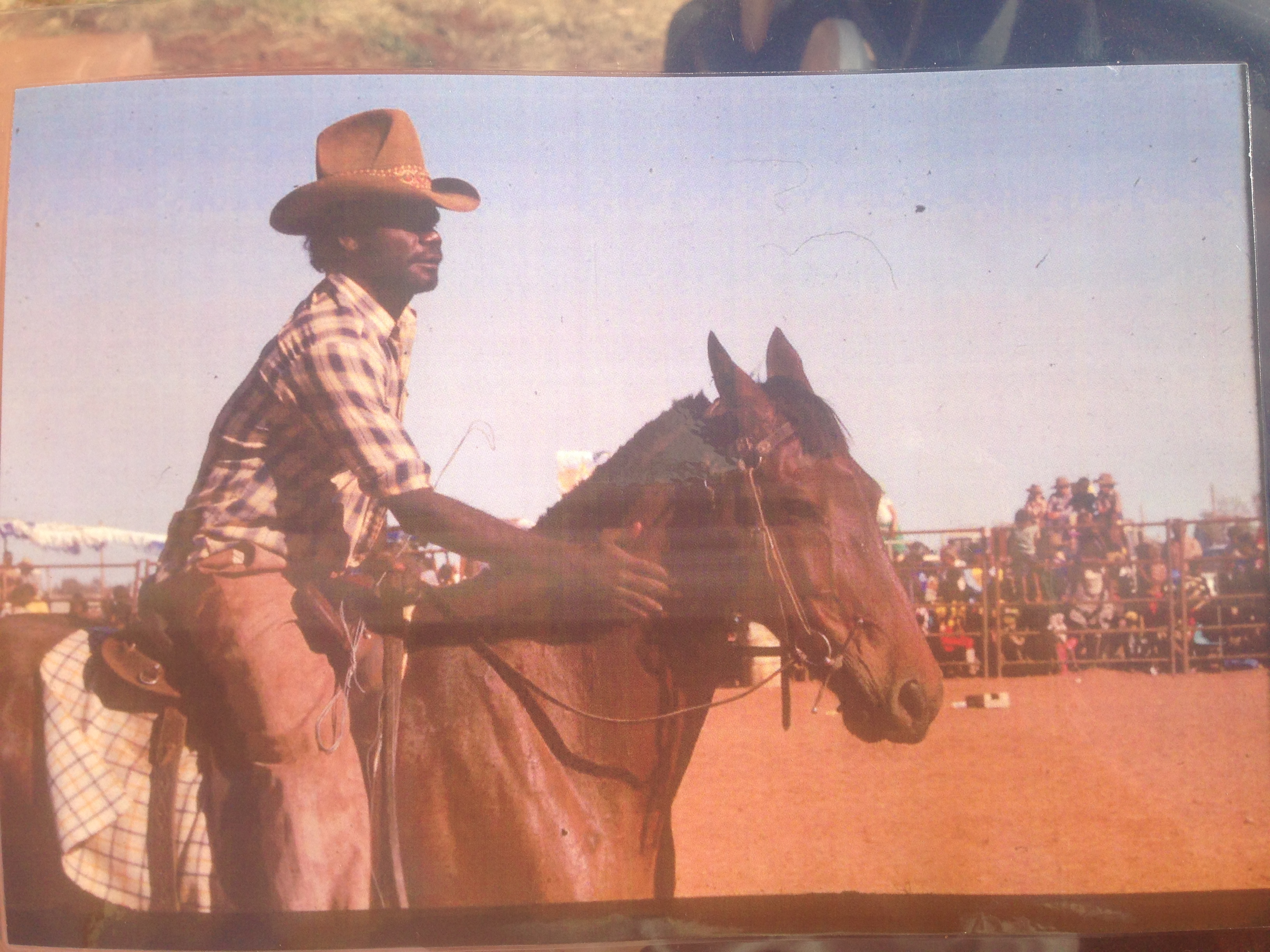 Image of Mark as a young man on a brown horse, patting its neck in a rodeo with people watching from the gates.