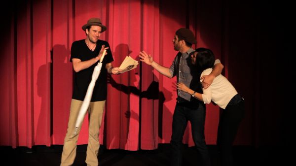 Three people on a stage in front of a red curtain. In the center a man with a hat and umbrella has his hands off, warding off the couple fro the right who are reaching emploringly toward him.