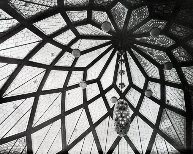 The exhibit features Bruno Taut’s Glashaus interior cupola from 1914, showcasing an intricately designed, flower-like structure with central lighting suspended from the ceiling’s tip or core.