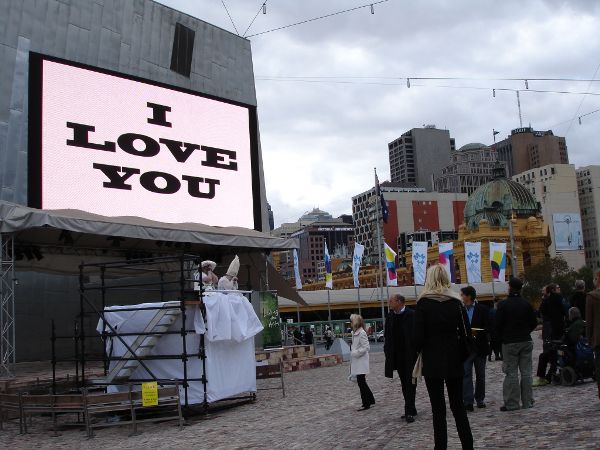 Two street performers, dressed in white, perform on scaffolding beneath a digital screen on a street corner with passersby looking on.