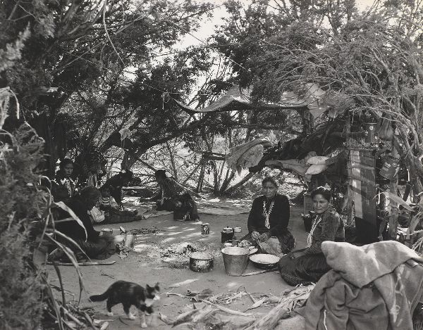 Nine family members, women and children, sit inside a shelter in glade of trees, engaged in domestic activities, with a cat in the foreground.