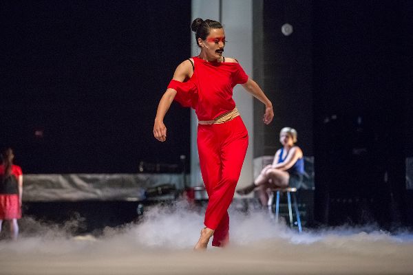 A dancer stands twisting, arms out, hips to side, one leg crossed. She wears her hair in a bun, and a mustache. Far behind, a woman sits on a chair.