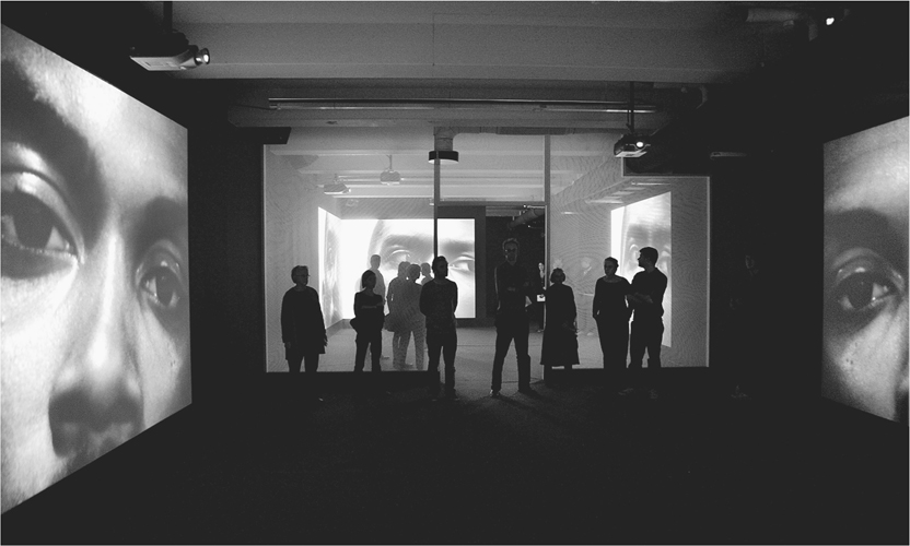 A photograph of a gallery with environmental artwork with projectors on the walls and people staring at them.